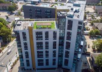 Aerial view of a modern, multi-story apartment building with a rooftop green space and tennis court, surrounded by streets, parked cars, and trees in an urban area.