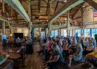 A group of people sit attentively on chairs inside a rustic, wooden-beamed hall with American flags, plants, and natural light streaming through large windows. Two people stand at the front, appearing to address the audience.