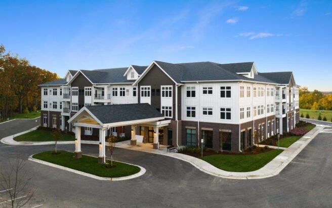 A modern, three-story building with white siding and brown brick accents, featuring large windows and a covered entrance, surrounded by a parking lot and green landscaping under a clear blue sky.