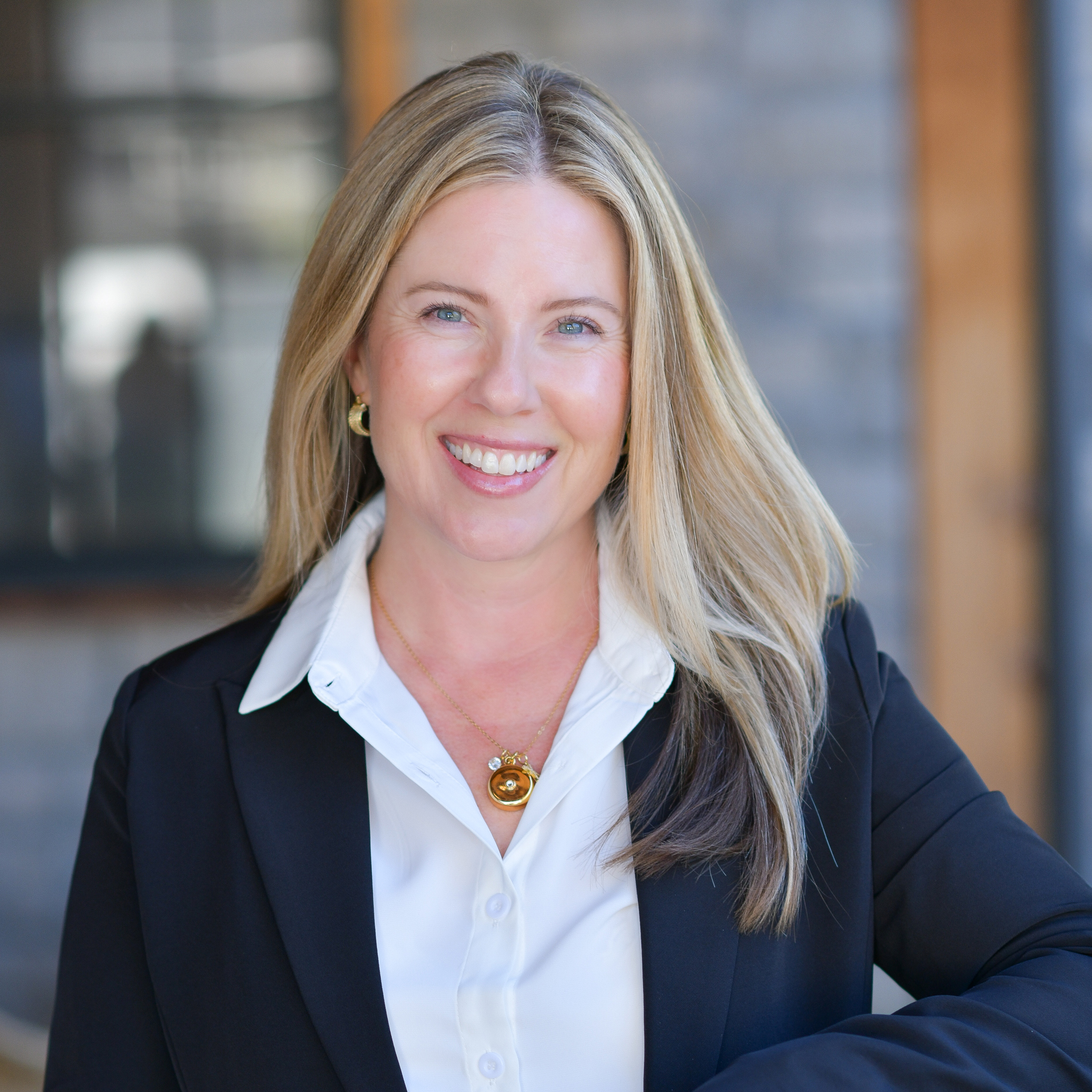 A woman with long blonde hair, wearing a black blazer over a white shirt and a gold necklace, smiles while standing in front of a blurred, modern building background.