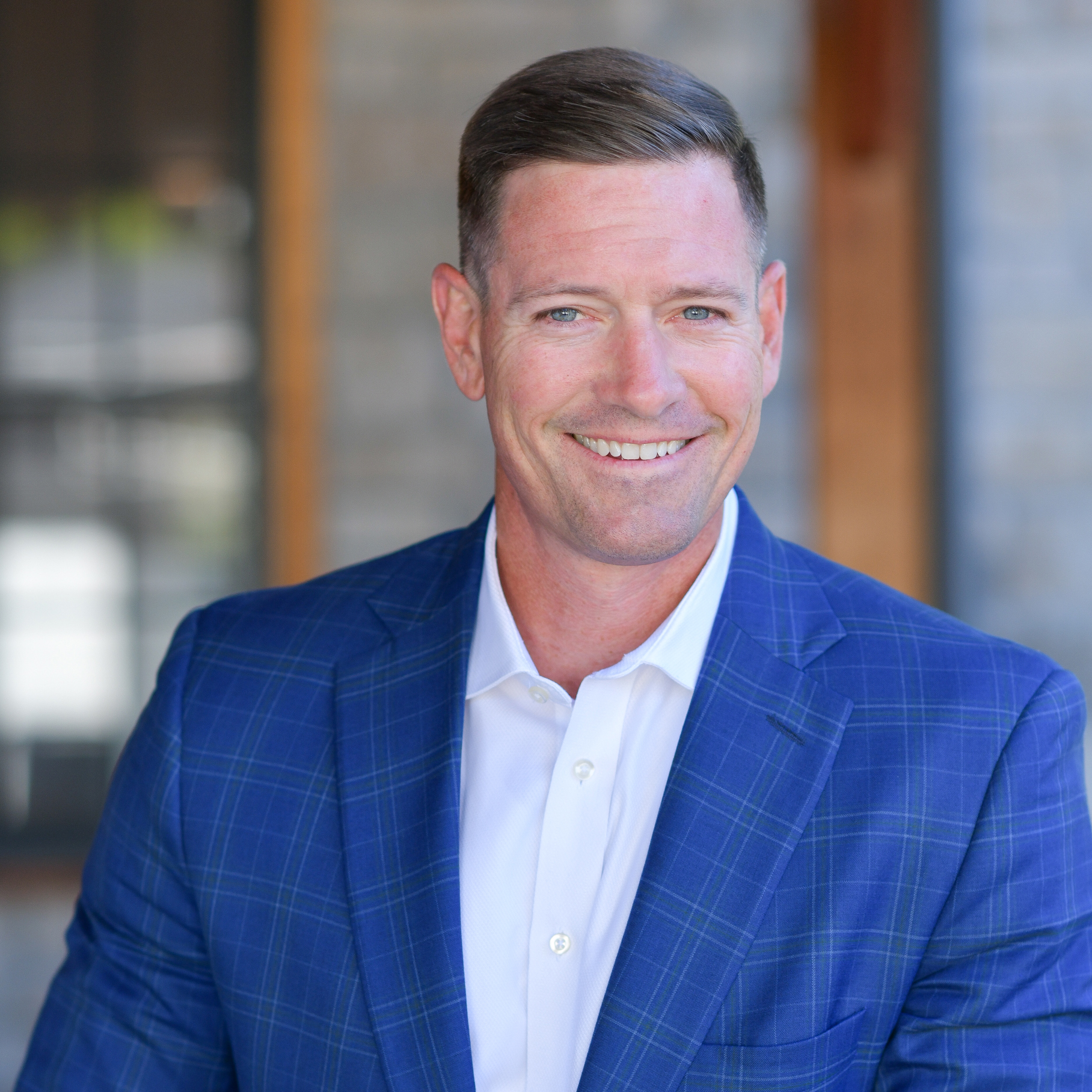 A man in a blue checked suit jacket and white shirt smiles while posing in front of a blurred brick and glass background.