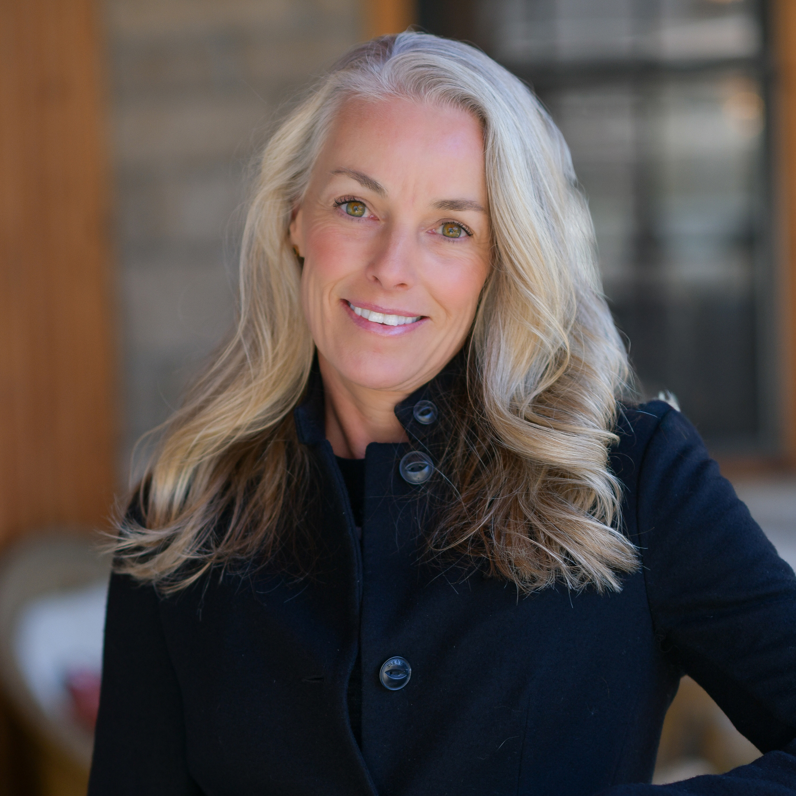 A woman with long, wavy gray-blonde hair and a black coat smiles outdoors, standing in front of a blurred background with a window and wooden siding.