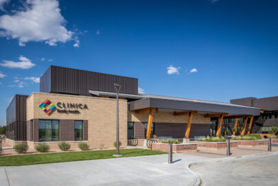 A modern medical clinic building with a tan brick exterior, large windows, and a shaded entrance supported by wooden beams, under a blue sky with scattered clouds. The sign reads CLINICA Family Health.