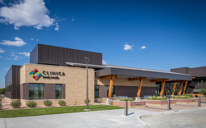 A modern medical clinic building with a tan brick exterior, large windows, and a shaded entrance supported by wooden beams, under a blue sky with scattered clouds. The sign reads CLINICA Family Health.