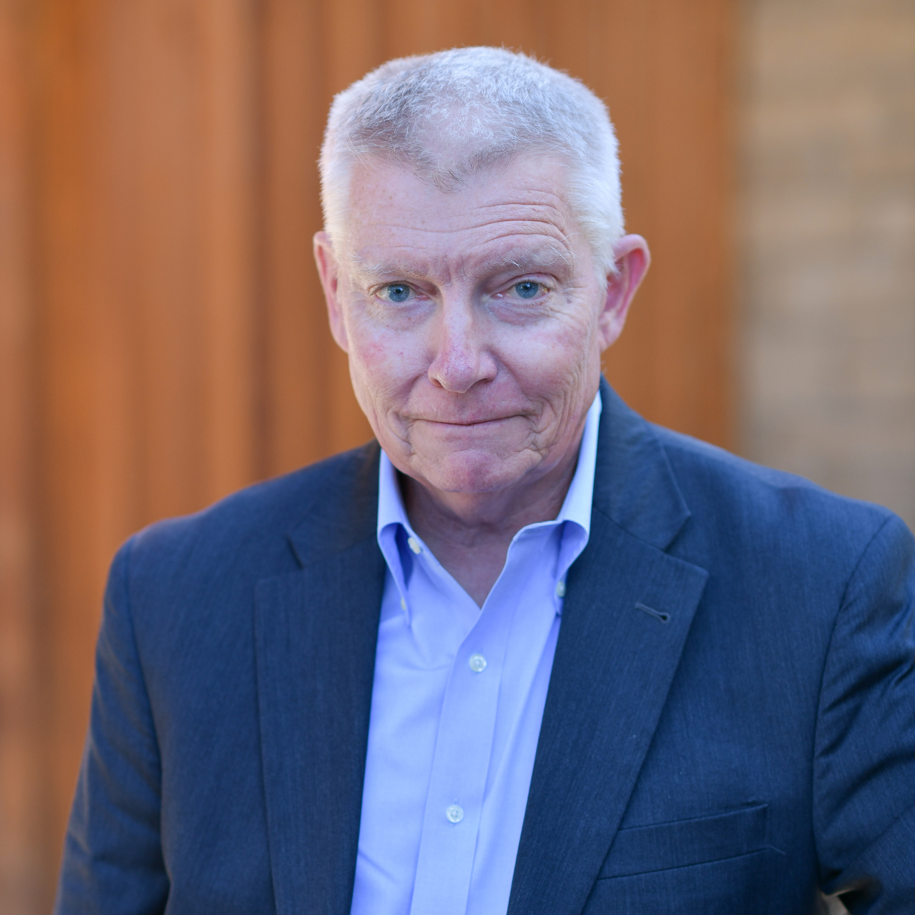 A senior man with short gray hair, wearing a blue suit jacket and light purple shirt, stands outdoors in front of a wooden background, looking at the camera with a slight smile.