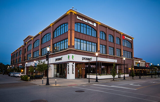A three-story brick building at dusk with large windows. Signs for Brown Smith Wallace and PNC Bank are visible on the upper floors. The ground floor features Prasino restaurant with outdoor seating.