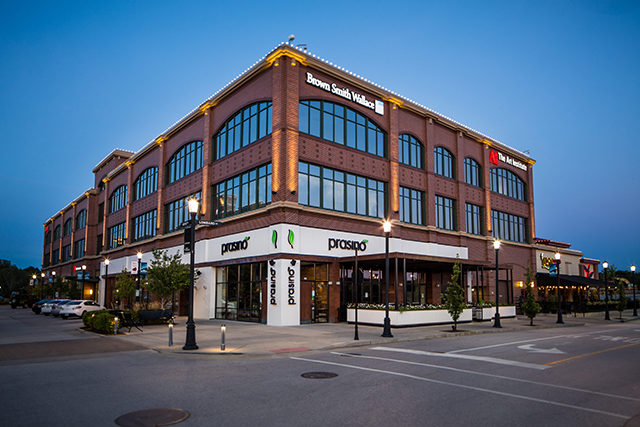 A three-story brick building at dusk with large windows. Signs for Brown Smith Wallace and PNC Bank are visible on the upper floors. The ground floor features Prasino restaurant with outdoor seating.