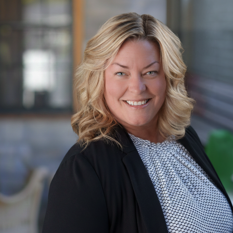 A woman with blonde, wavy hair smiles and stands outdoors. She is wearing a black blazer over a patterned white blouse. The background is softly blurred, showing a building and some greenery.