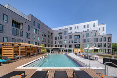 Modern apartment complex with gray brick exterior, large windows, and balconies. In the foreground is a swimming pool with lounge chairs, umbrellas, and a wooden pergola under a clear blue sky.