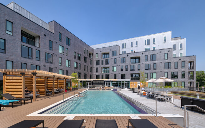 Modern apartment complex with gray brick exterior, large windows, and balconies. In the foreground is a swimming pool with lounge chairs, umbrellas, and a wooden pergola under a clear blue sky.