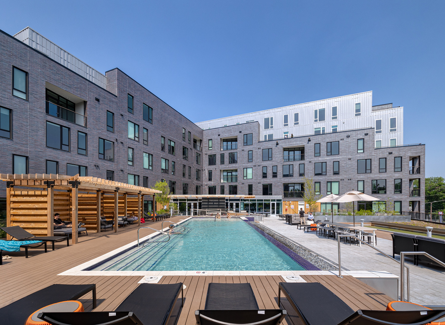 Modern apartment complex with gray brick exterior, large windows, and balconies. In the foreground is a swimming pool with lounge chairs, umbrellas, and a wooden pergola under a clear blue sky.