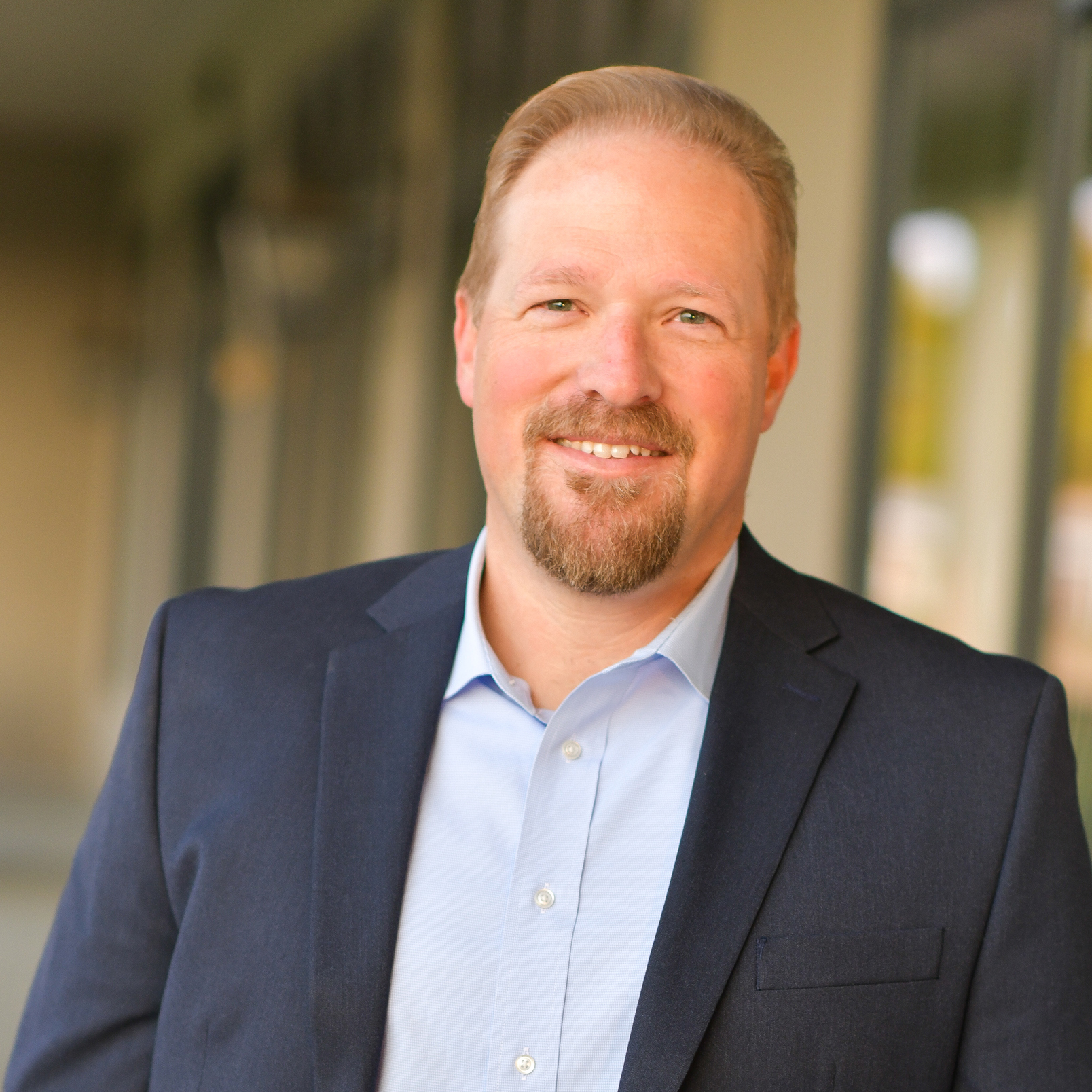 A smiling man with short blond hair and a beard wears a dark blue blazer and light blue shirt, standing in front of a blurred outdoor background with windows and columns.