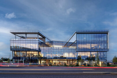 A modern glass office building with angular architecture is lit from within at dusk, reflecting the blue sky. Light trails from passing cars are visible on the road in the foreground.
