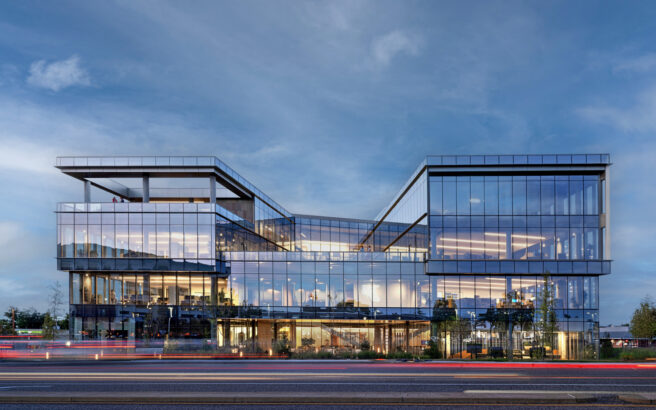 A modern glass office building with angular architecture is lit from within at dusk, reflecting the blue sky. Light trails from passing cars are visible on the road in the foreground.