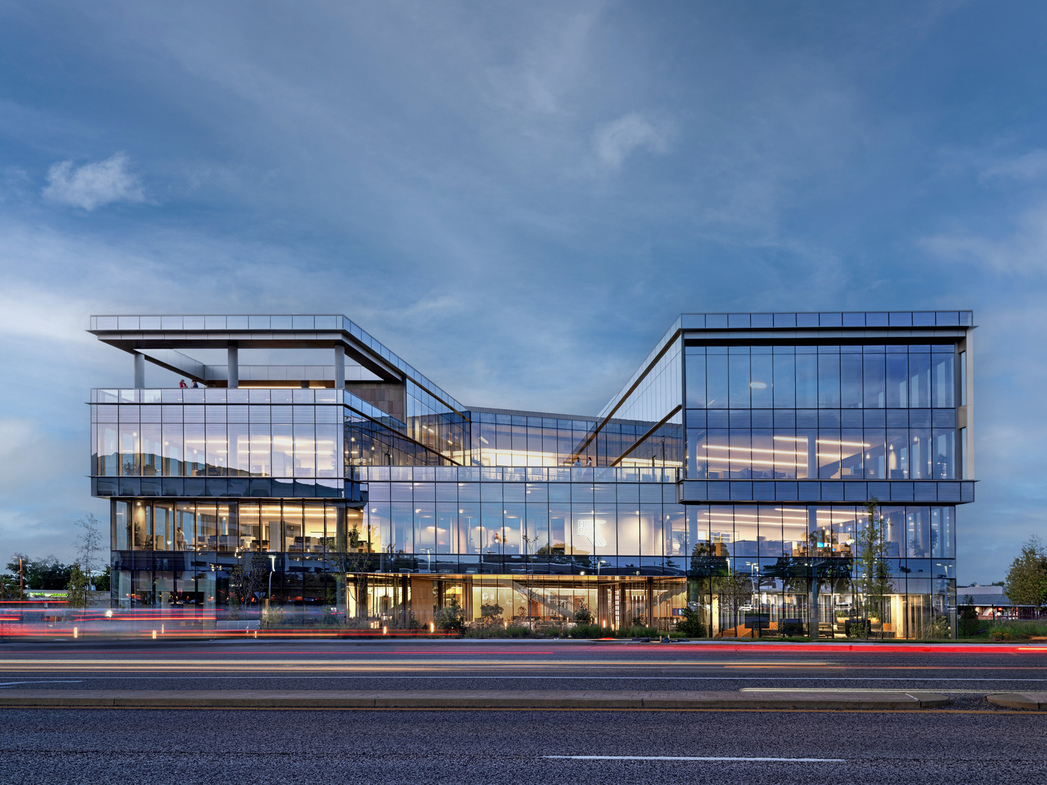 A modern glass office building with angular architecture is lit from within at dusk, reflecting the blue sky. Light trails from passing cars are visible on the road in the foreground.