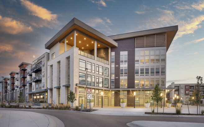 Modern, multi-story apartment building with large windows, balconies, and prominent geometric architecture, lit by warm exterior lights, under a colorful evening sky. Landscaping and a curved driveway surround the entrance.