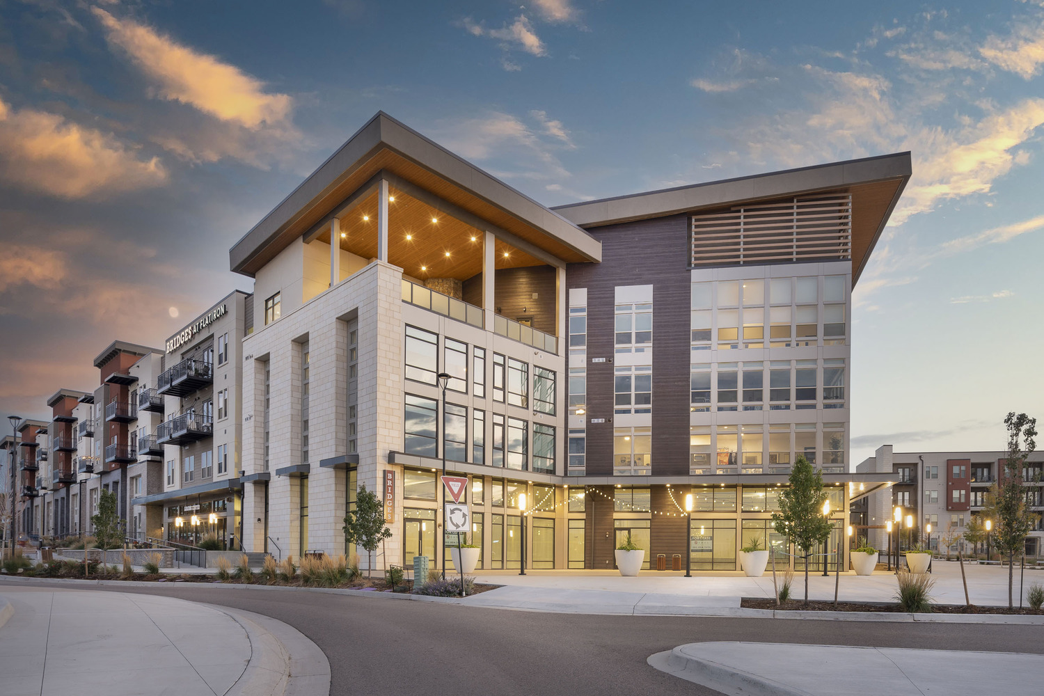 Modern, multi-story apartment building with large windows, balconies, and prominent geometric architecture, lit by warm exterior lights, under a colorful evening sky. Landscaping and a curved driveway surround the entrance.