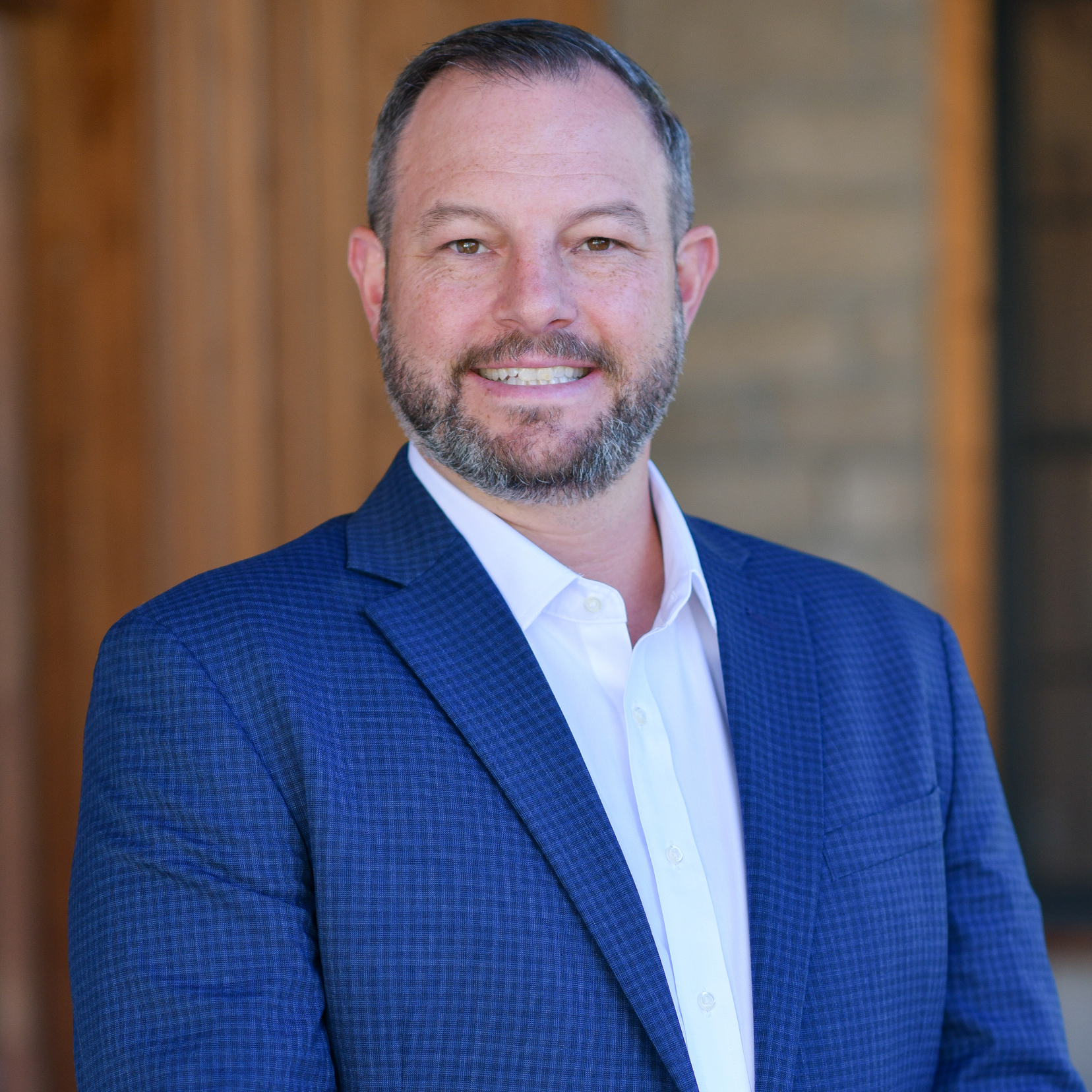 A man with short hair and a beard, wearing a blue checked suit jacket and white shirt, smiles at the camera while standing in front of a blurred outdoor background.
