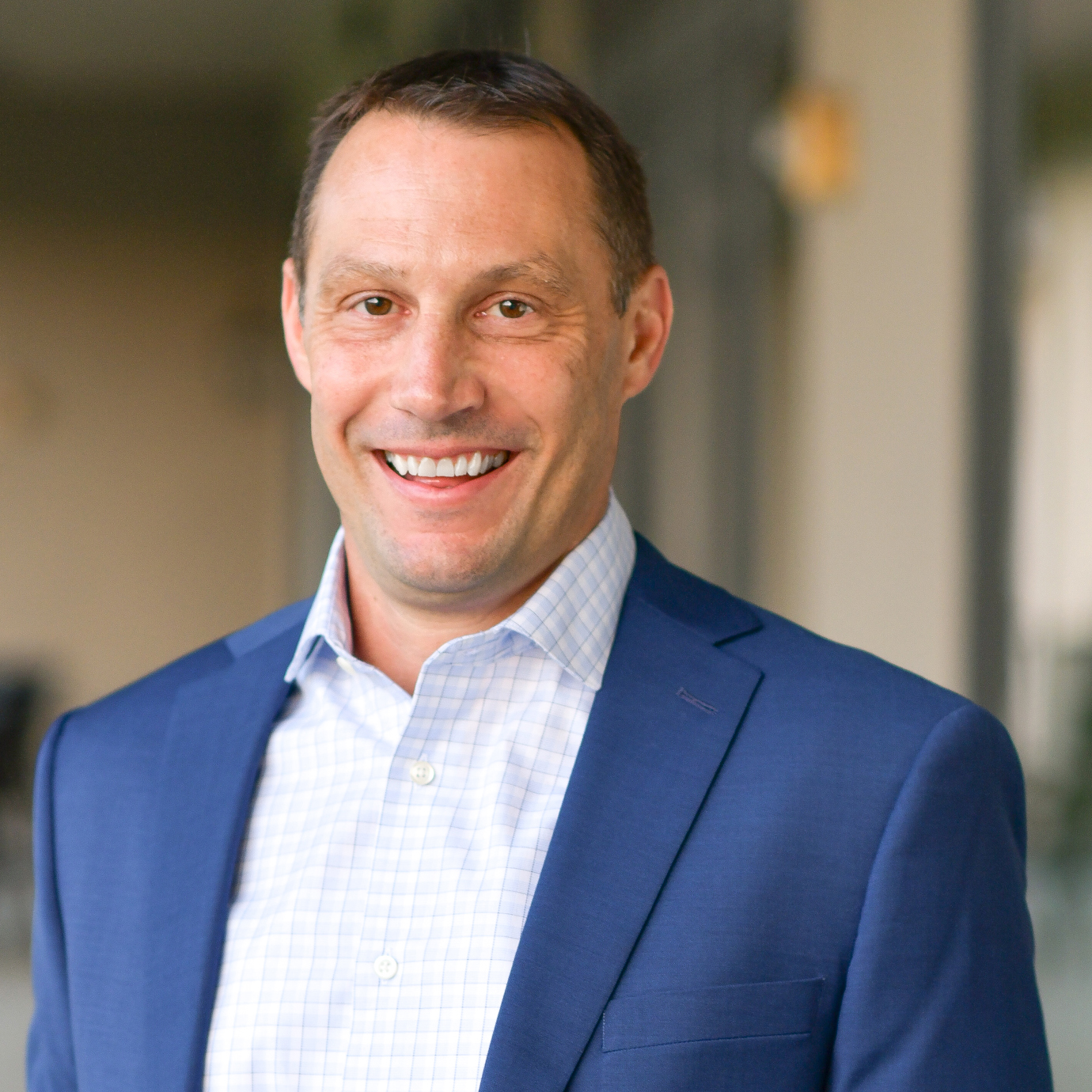 A man in a blue suit and checkered white shirt smiles at the camera while standing outdoors in a blurred background.