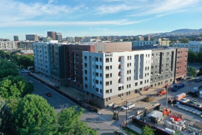 Aerial view of a multi-story apartment building under construction in an urban area, surrounded by trees, roads, cranes, and construction equipment, with a city skyline and mountains in the background.