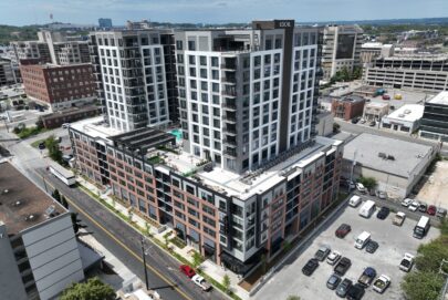 Aerial view of a modern multi-story apartment building with balconies, a rooftop pool, and surrounding city streets, cars parked in nearby lots, and other urban buildings in the background under a partly cloudy sky.