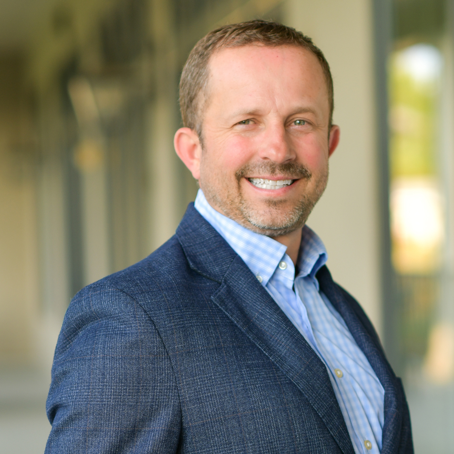 Smiling man with short hair and beard wearing a blue suit jacket and checked shirt, standing in front of a blurred outdoor background.