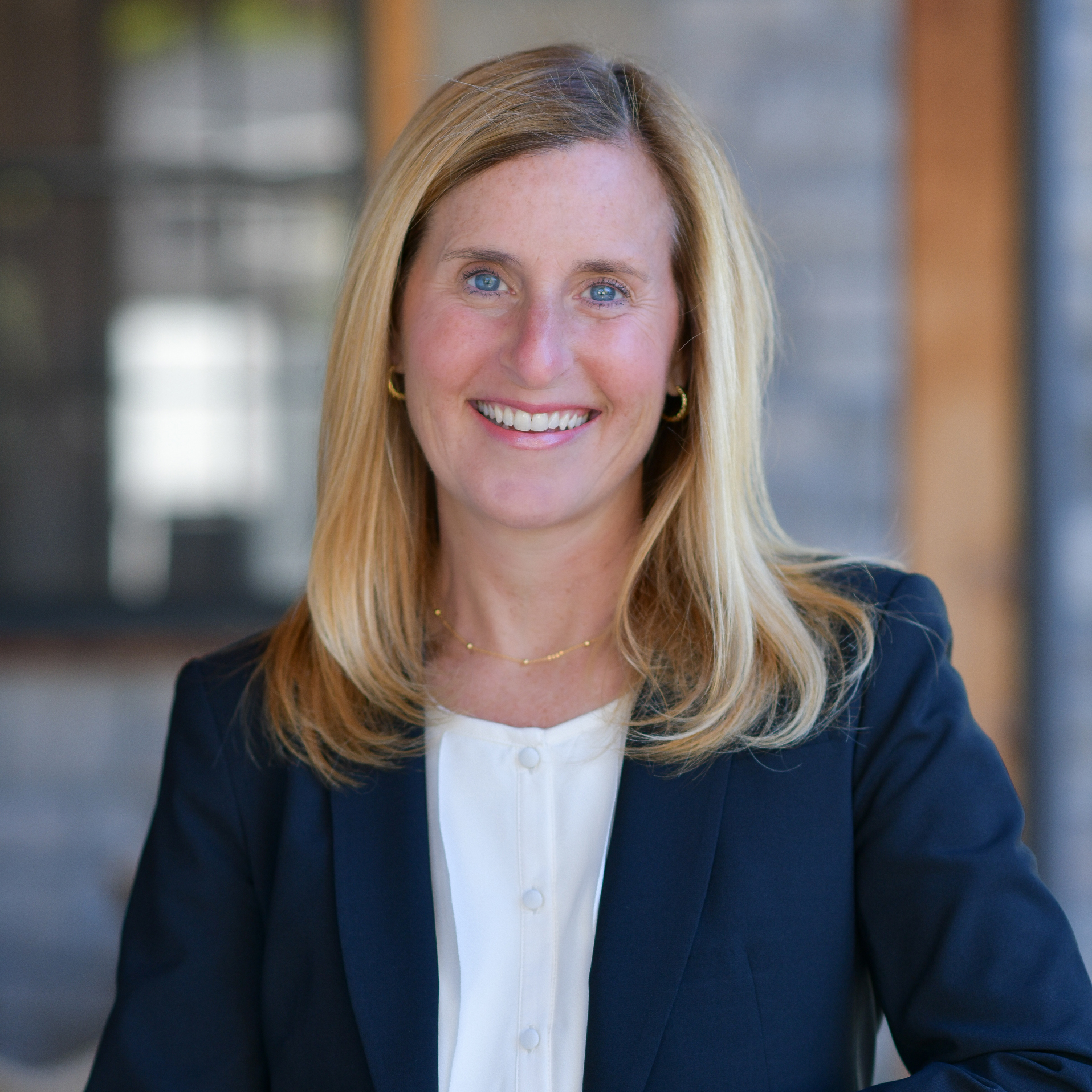 A woman with blonde hair, wearing a navy blazer and white blouse, smiles at the camera. She stands in front of a blurred background with a window and wooden beams.