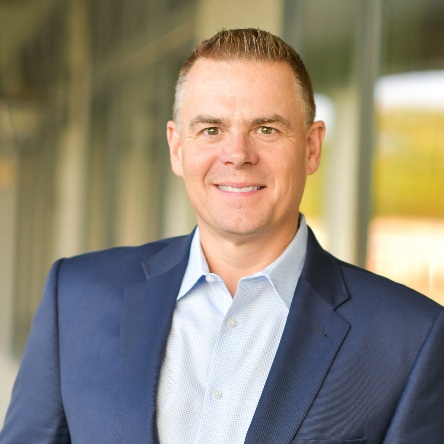 A man with short light brown hair, wearing a blue blazer and light blue collared shirt, smiles at the camera while standing indoors in front of large windows.