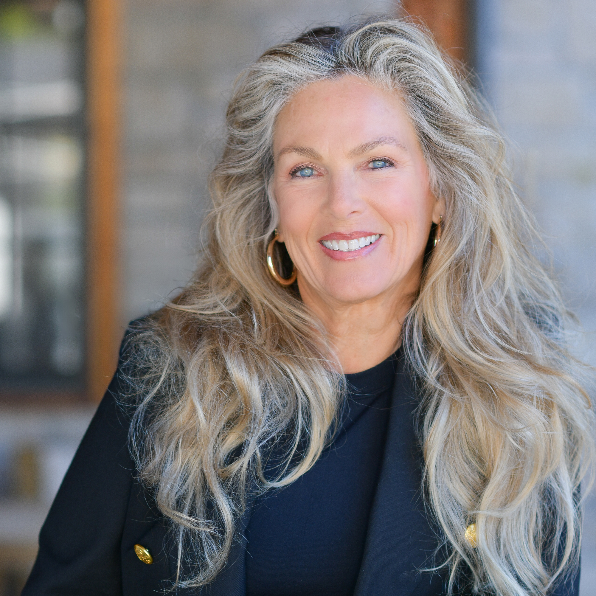 A smiling woman with long, wavy blonde hair wearing a black outfit and gold hoop earrings stands in front of a blurred background.
