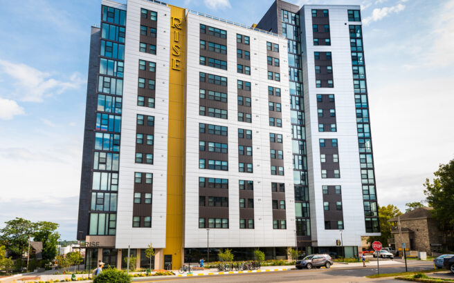 A tall, modern apartment building with white and black panels and a yellow vertical accent reading RISE. The building has many windows and is surrounded by trees, greenery, and a few parked bicycles.