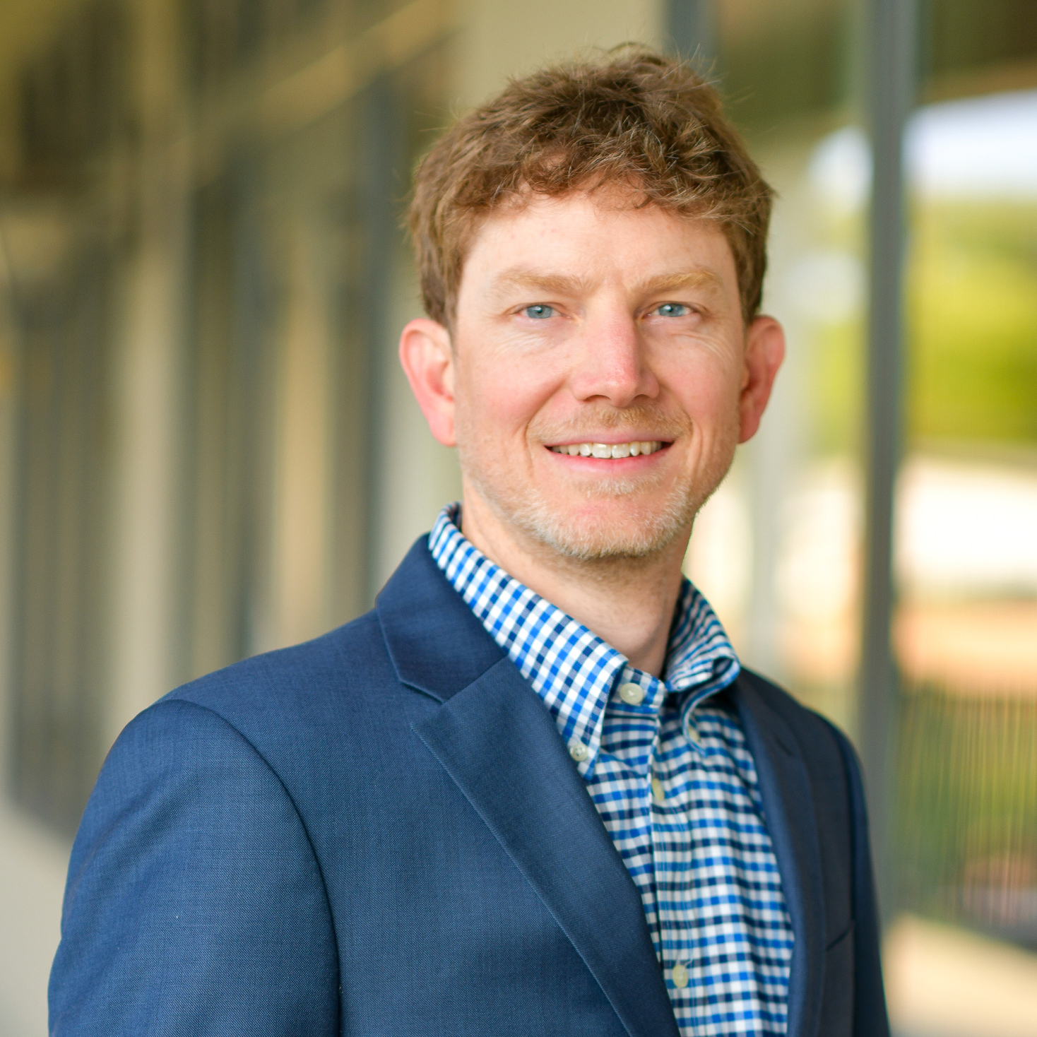 A man with wavy brown hair and a short beard, wearing a blue blazer and a blue checkered shirt, is smiling while standing in front of a blurred glass and concrete background.