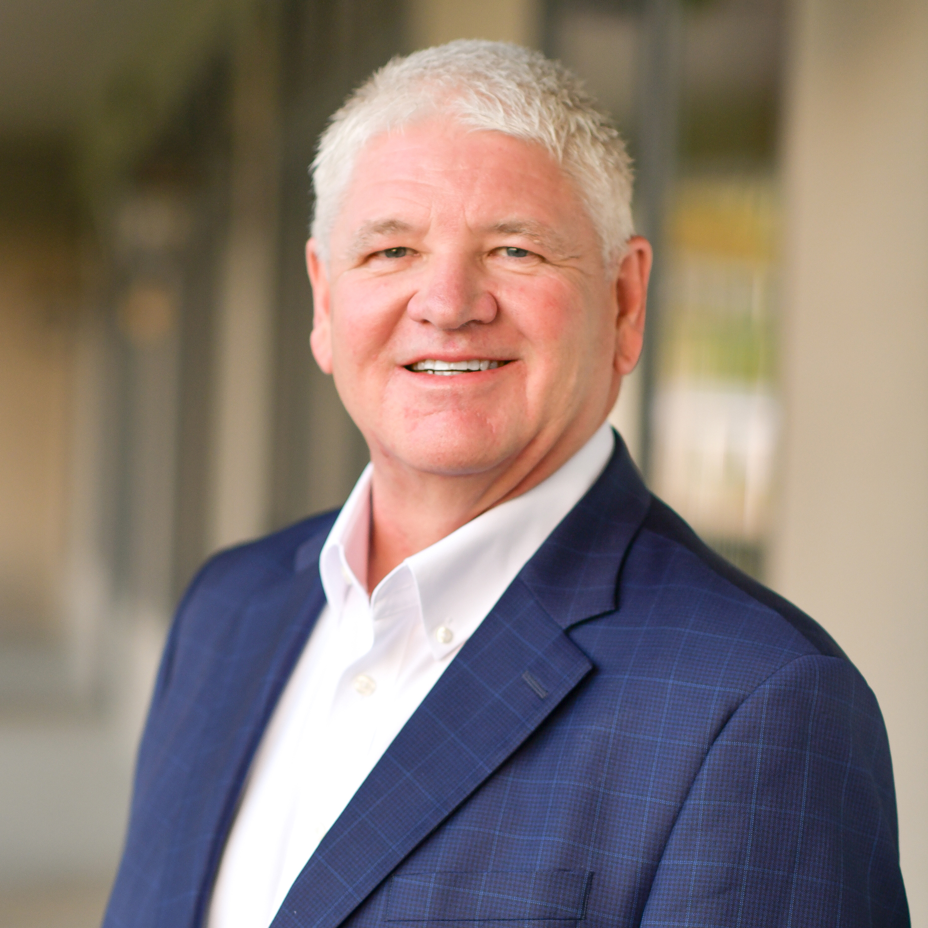 A smiling older man with short white hair wears a blue checked blazer over a white dress shirt. He is standing outdoors in front of a blurred building background.