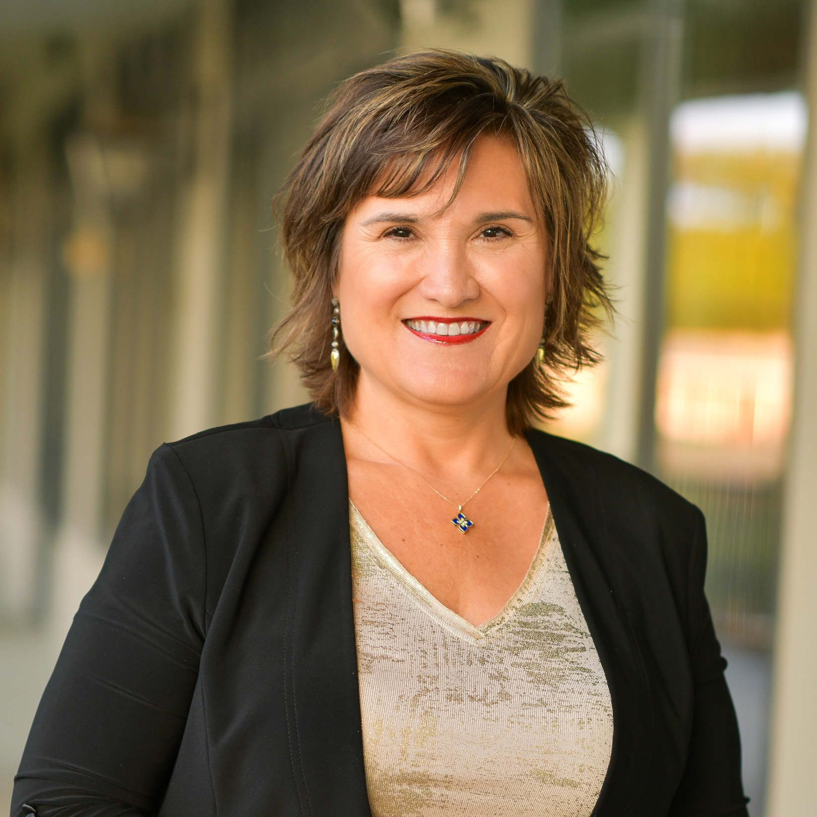 A woman with short brown hair, wearing a black blazer over a beige top and a pendant necklace, smiles at the camera outdoors with a blurred background.
