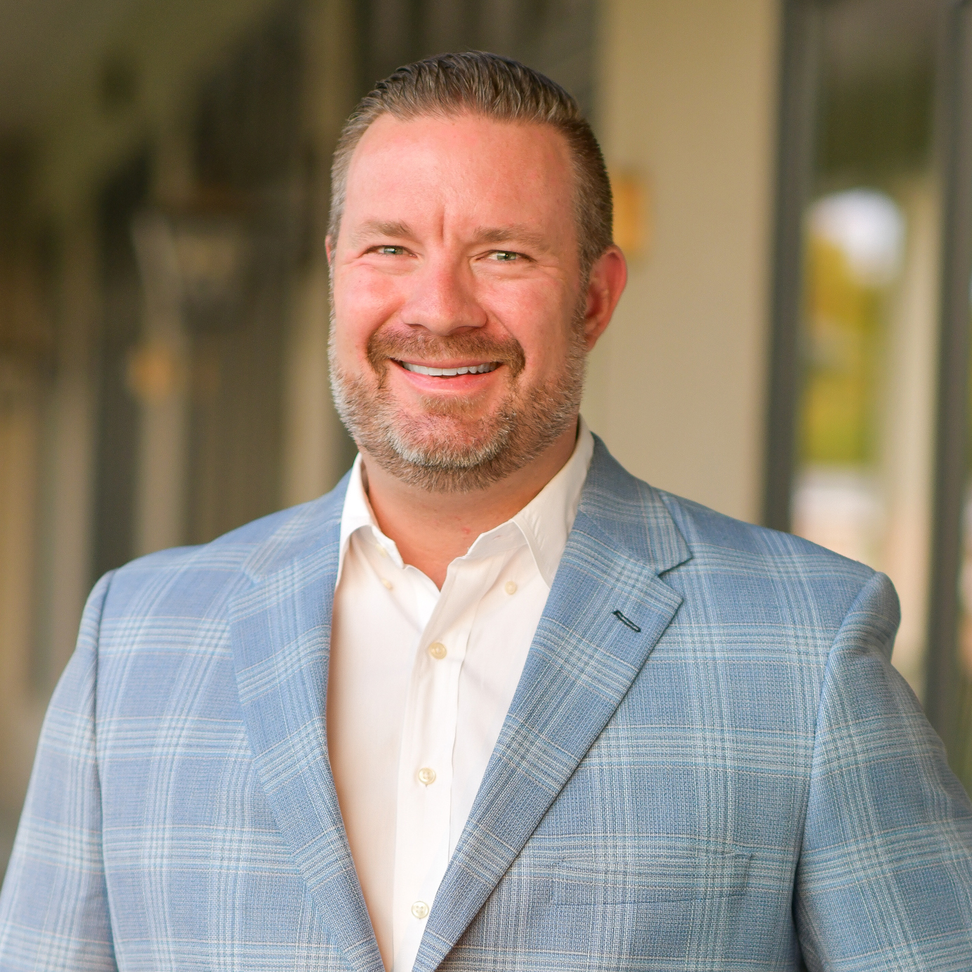 A smiling man with short hair and a beard, wearing a light blue plaid suit jacket and white shirt, stands outside in front of a building with large windows.