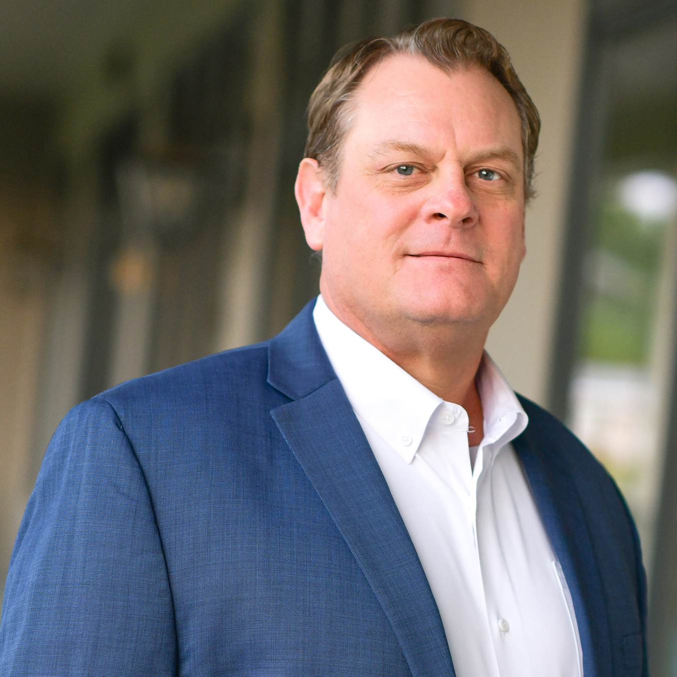 A man in a blue suit jacket and white shirt stands outdoors, looking confidently at the camera. The background is blurred with neutral tones.