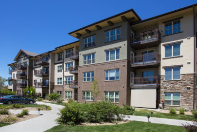 A modern four-story apartment building features brick, stone, and stucco siding, with balconies and large windows. A few parked cars and landscaped greenery are visible in front under a clear blue sky.