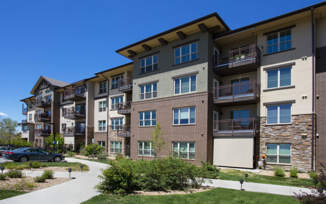 A modern four-story apartment building features brick, stone, and stucco siding, with balconies and large windows. A few parked cars and landscaped greenery are visible in front under a clear blue sky.