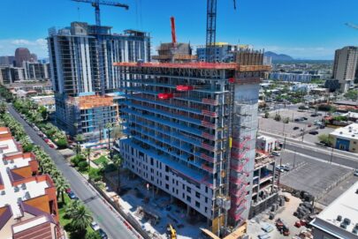 Aerial view of a mid-rise building under construction in a city, surrounded by cranes, nearby buildings, roads, and parked cars under a clear blue sky.