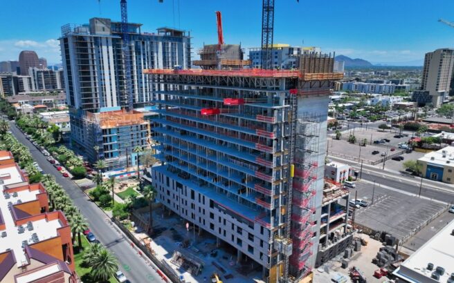 Aerial view of a mid-rise building under construction in a city, surrounded by cranes, nearby buildings, roads, and parked cars under a clear blue sky.