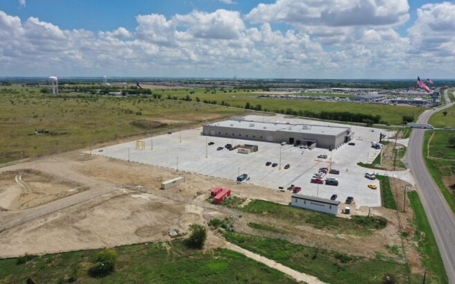 Aerial view of a large industrial building with a spacious parking lot, surrounded by grassy fields and dirt areas, with a road nearby and a few vehicles parked outside under a partly cloudy sky.