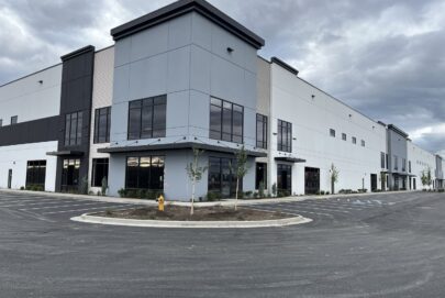 A large, modern industrial or warehouse building with tall windows, gray and white exterior walls, and an empty parking lot under a cloudy sky. Young trees and a yellow fire hydrant are near the entrance.