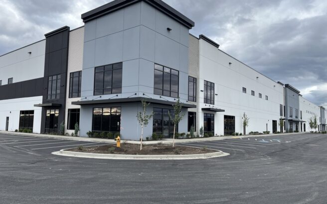 A large, modern industrial or warehouse building with tall windows, gray and white exterior walls, and an empty parking lot under a cloudy sky. Young trees and a yellow fire hydrant are near the entrance.