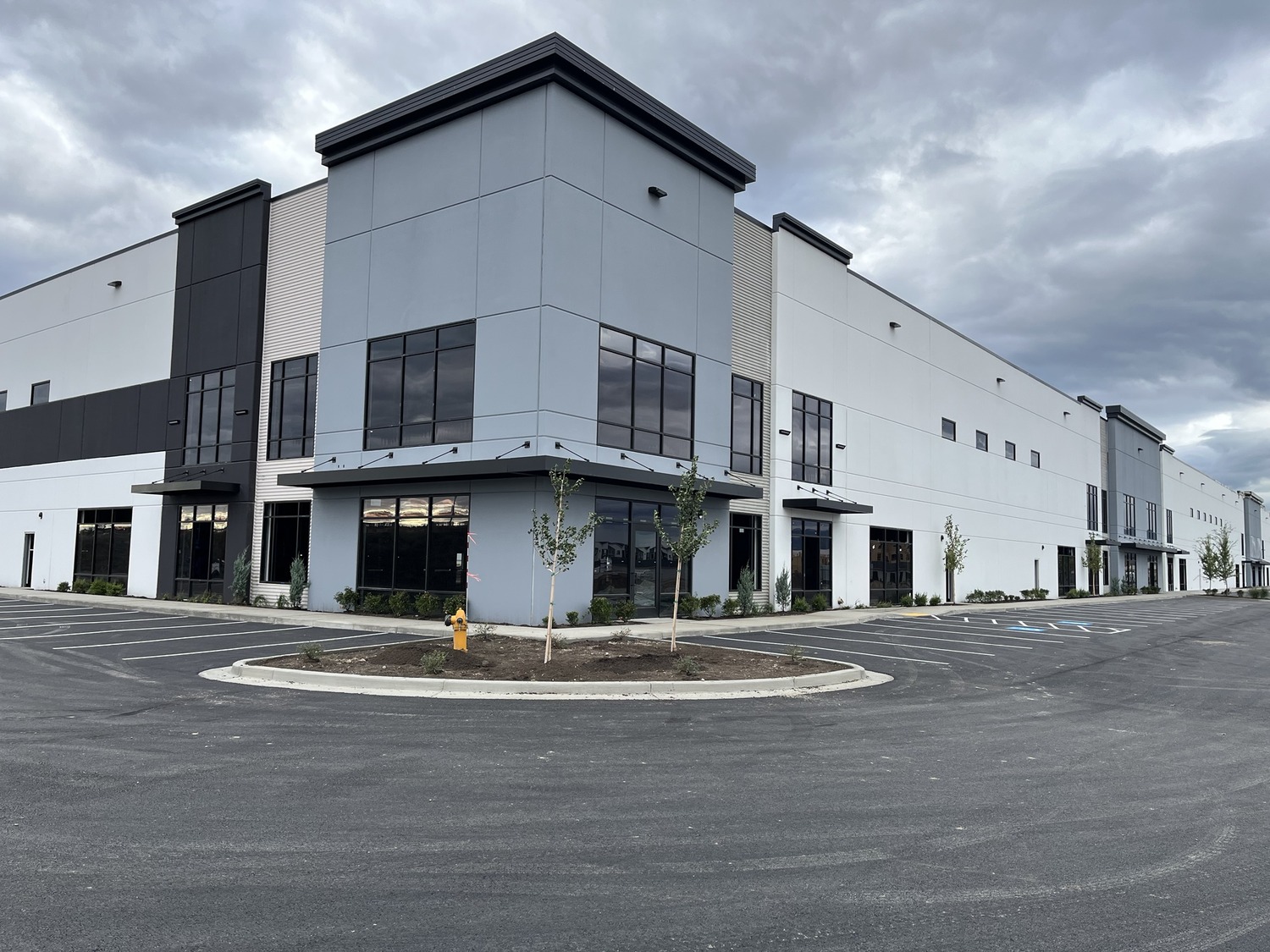 A large, modern industrial or warehouse building with tall windows, gray and white exterior walls, and an empty parking lot under a cloudy sky. Young trees and a yellow fire hydrant are near the entrance.