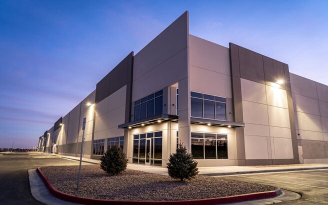 A modern industrial warehouse building with large windows and exterior lights illuminating the facade at dusk. The parking lot is empty, and small shrubs are planted along the sidewalk.
