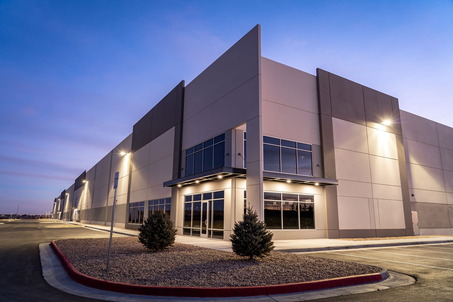 A modern industrial warehouse building with large windows and exterior lights illuminating the facade at dusk. The parking lot is empty, and small shrubs are planted along the sidewalk.