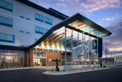 Modern hotel exterior with a colorful, illuminated canopy, large “abl” signage, and glass entrance. The building has sleek lines, large windows, and is shown at dusk with a dramatic sky in the background.