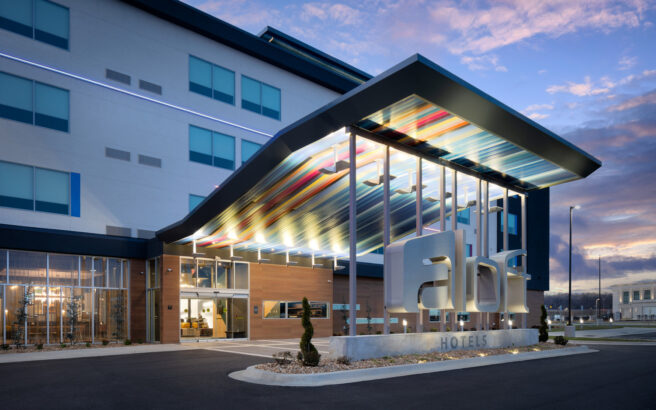 Modern hotel exterior with a colorful, illuminated canopy, large “abl” signage, and glass entrance. The building has sleek lines, large windows, and is shown at dusk with a dramatic sky in the background.