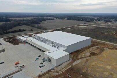 Aerial view of a large, white-roofed industrial warehouse surrounded by dirt, construction vehicles, and partially developed land, with fields and forested areas in the background under a cloudy sky.