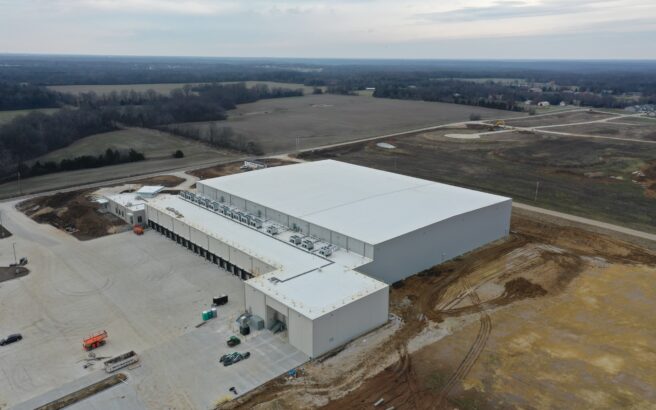Aerial view of a large, white-roofed industrial warehouse surrounded by dirt, construction vehicles, and partially developed land, with fields and forested areas in the background under a cloudy sky.