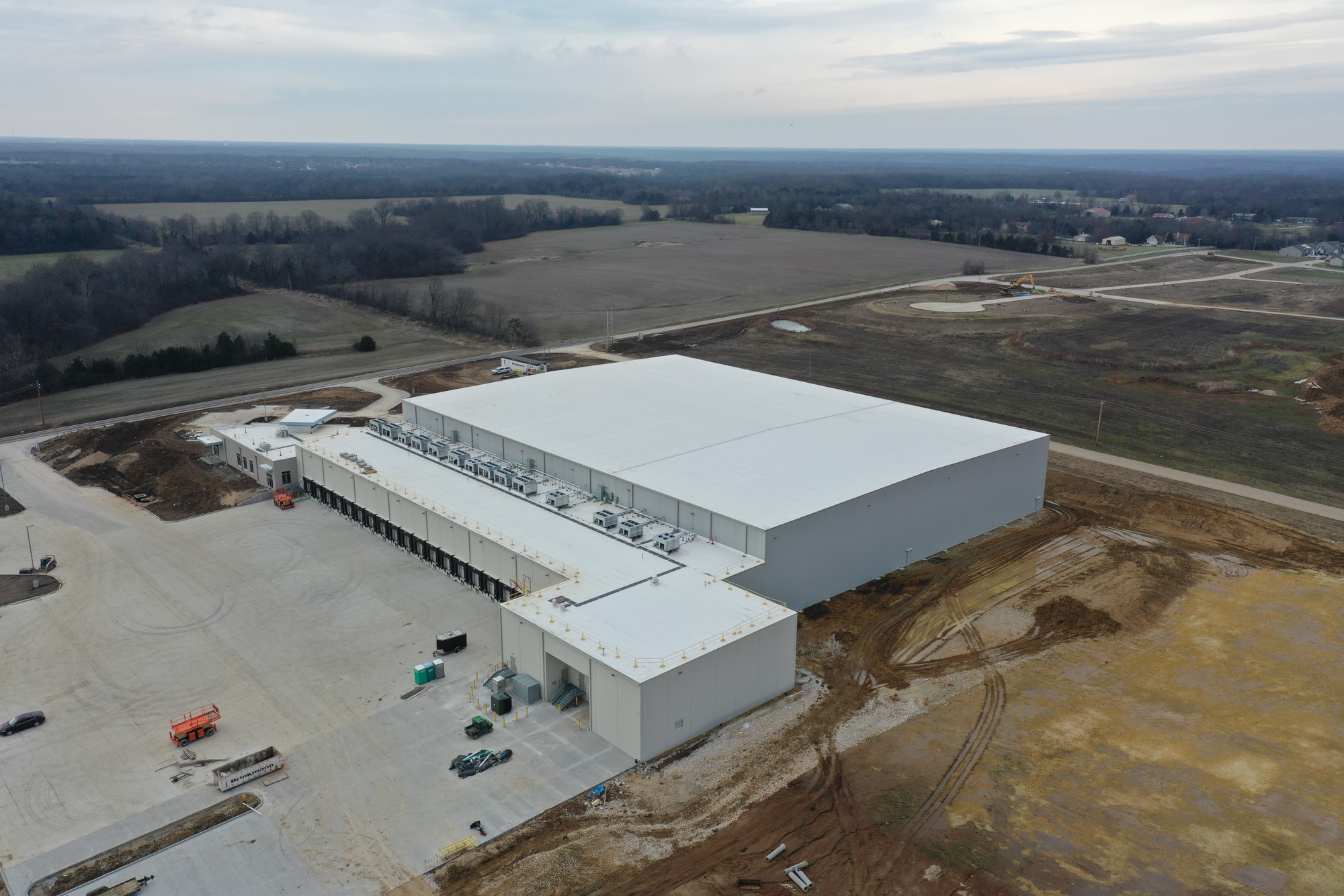 Aerial view of a large, white-roofed industrial warehouse surrounded by dirt, construction vehicles, and partially developed land, with fields and forested areas in the background under a cloudy sky.